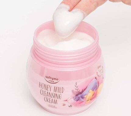 Fingers scooping white cleansing cream from a pink jar of Softymo Honey Mild Cleansing Cream featuring Winnie the Pooh.