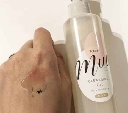 Hand with makeup smudge being cleansed by a drop of Kracie Muo Cleansing Oil, with the bottle visible.